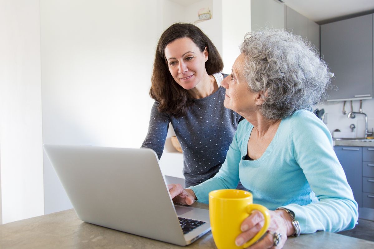 Excited senior woman consulting daughter on using online service. Young and elderly women using laptop in kitchen. Technical support concept